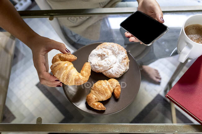 Mujer usando smartphone con croissant y taza de café en mesa de madera - foto de stock