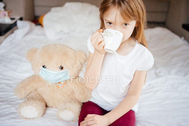 Fille boit du thé avec un ours en peluche dans un masque médical sur un lit blanc — Photo de stock