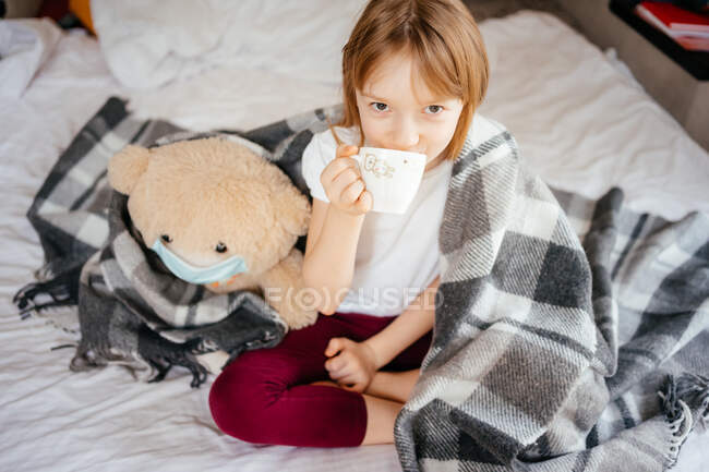 Menina bebe chá com um ursinho de pelúcia em uma máscara médica em uma cama branca — Fotografia de Stock