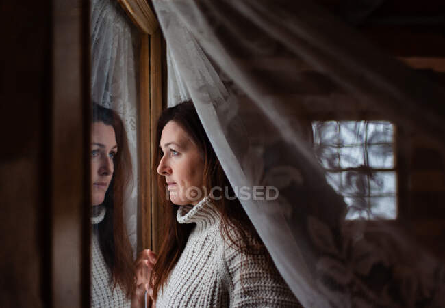 Attractive woman looking through window behind a lace curatin. — Stock Photo