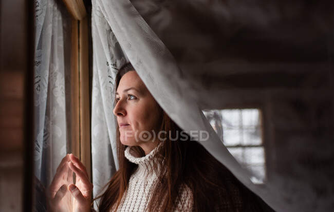 Приваблива жінка дивиться крізь вікно за мереживом куратан . — стокове фото