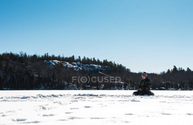 Niño pescando mientras está sentado en la nieve - foto de stock
