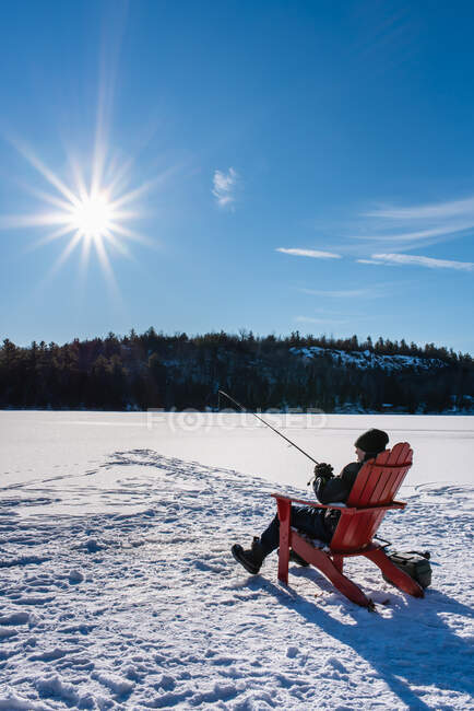 Niño pescando mientras está sentado en la nieve - foto de stock