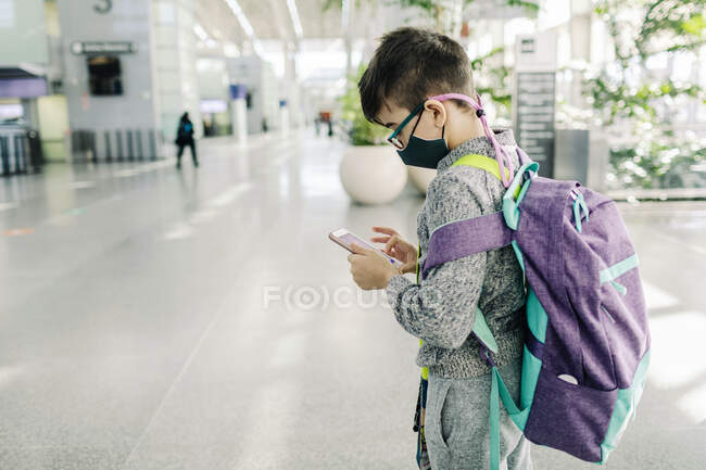 Niño preadolescente con máscara comprobando el teléfono en la terminal de llegadas del aeropuerto - foto de stock