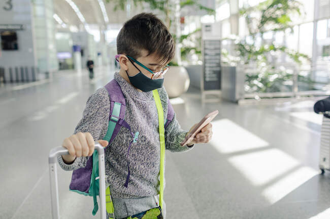 Pre-teen boy wearing mask checking phone in airport arrivals terminal — Stock Photo
