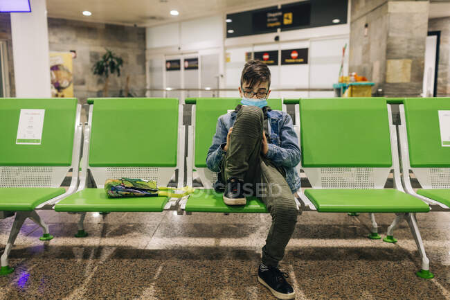 Pré-adolescent garçon portant un masque en utilisant le téléphone assis dans le terminal de l'aéroport — Photo de stock