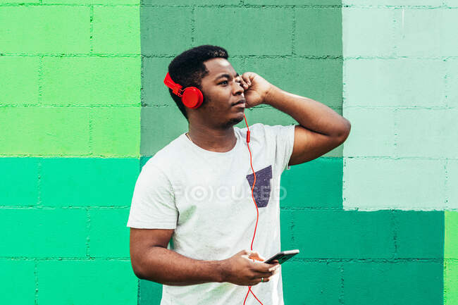 Afro garçon noir américain sur fond de mur vert. Écouter de la musique avec un casque et un téléphone portable. — Photo de stock