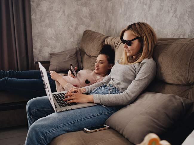 Dos mujeres jóvenes se sientan en el sofá con un portátil y un teléfono inteligente - foto de stock