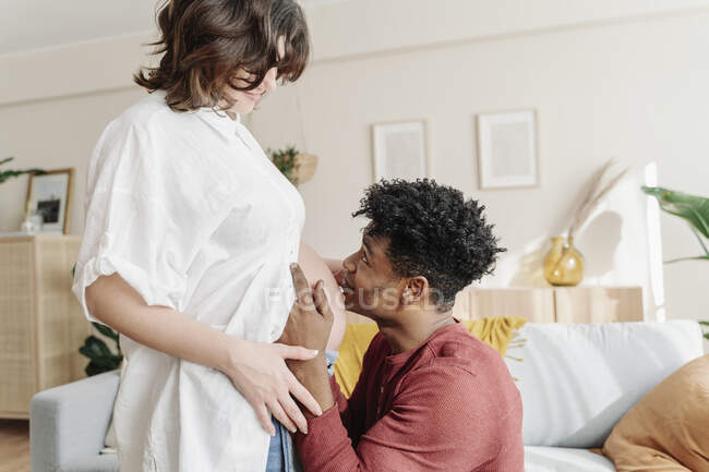 Side view of gentle African American happy male kissing belly of content pregnant female at home — Stock Photo