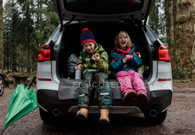 Irmãos sentados na bagageira de um carro comendo um lanche rindo juntos — Fotografia de Stock