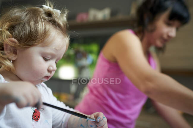 Menina bonito olhando para suas mãos pintadas com sua mãe no fundo — Fotografia de Stock