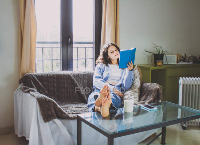 Молода жінка на дивані читає книжку з кавою. — стокове фото
