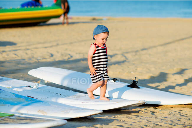 Miúdo bonito em uma praia andando descalço ao longo prancha windsurf — Fotografia de Stock