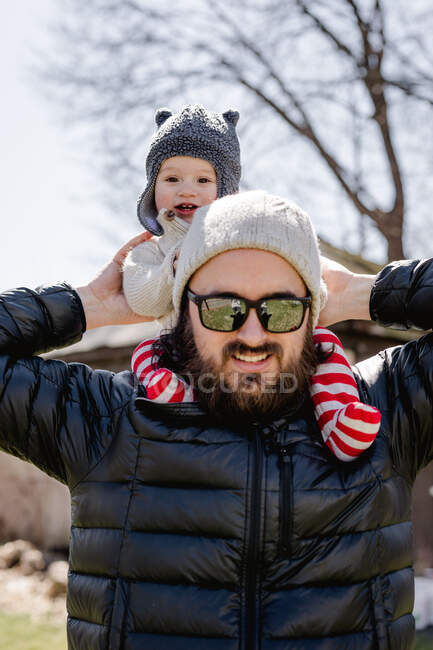 Bebé feliz en los hombros de papá en el patio trasero en la tarde fría - foto de stock