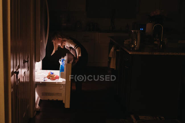 Tween girl looking into freezer drawer in dark kitchen — Stock Photo