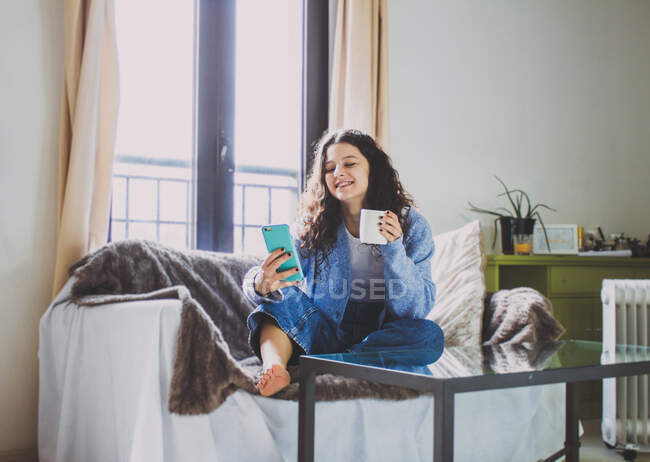 Jovem mulher tomando café da manhã no sofá com um café e celular — Fotografia de Stock