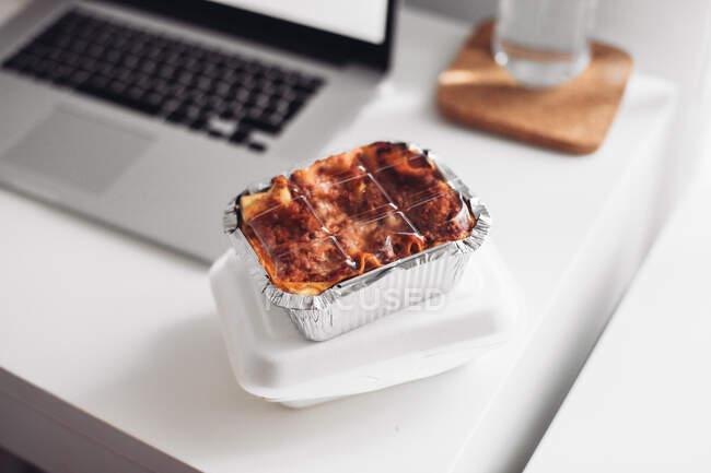 Une boîte à emporter avec des lasagnes, un ordinateur portable. Concept de manger à la maison — Photo de stock
