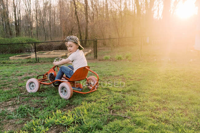 Молодий активний хлопчик з шапочкою в педальній машині на задньому дворі під час заходу сонця — стокове фото