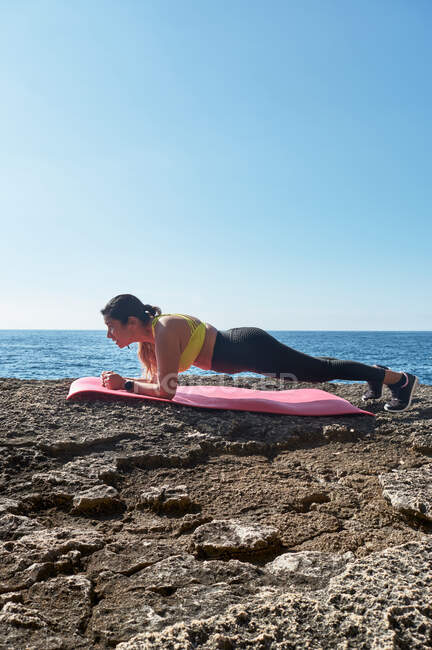 Latin woman, middle-aged, with yellow top, black leggings, training, doing physical exercises, plank, abs, climber's step, burning calories, keeping fit, outdoors by the sea,blue sky, day,sun — Stock Photo