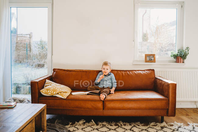 Adorable white blonde boy sitting on couch with pencil in his nose — Stock Photo
