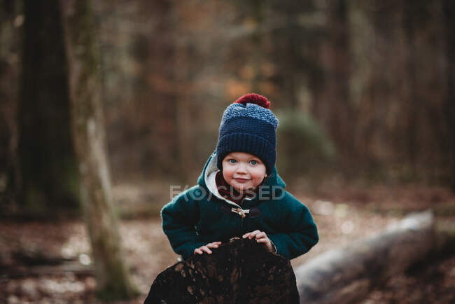 Чарівний молодий хлопчик лежить на колоді, посміхаючись у лісі в зимовому вовняному пальто — стокове фото