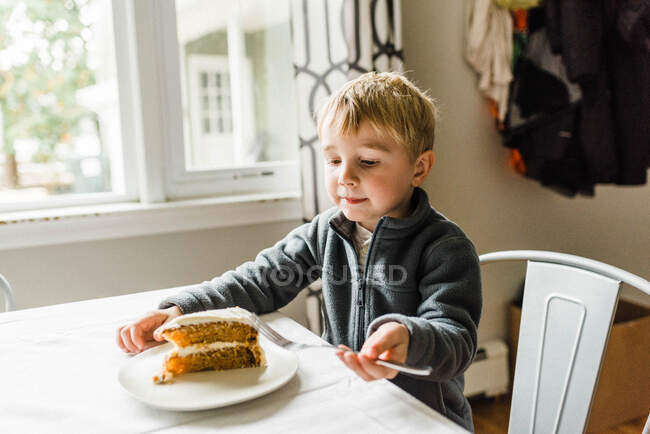 Pequeño chico rubio comiendo una rebanada de pastel de zanahoria casero en la mesa - foto de stock