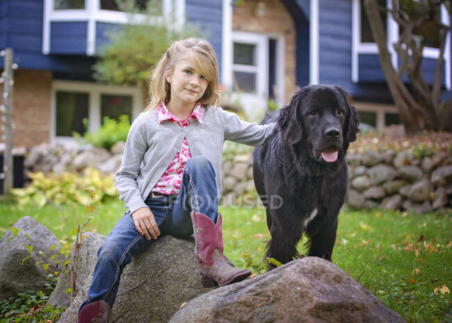 Jeune fille blonde assise dehors sur un rocher avec grand chien noir — Photo de stock