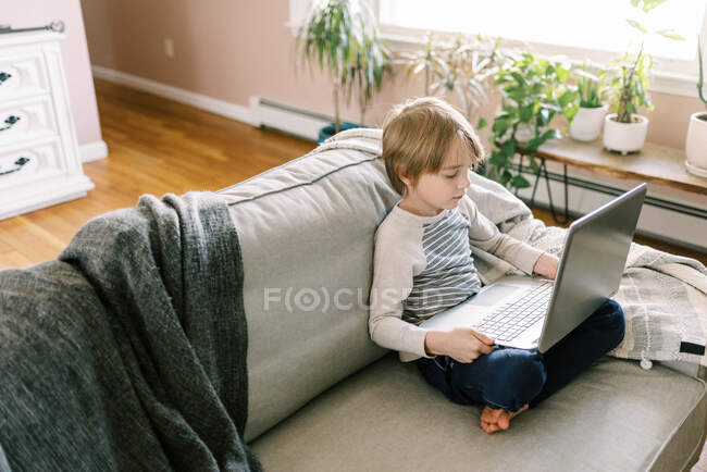 Niño haciendo la tarea de la escuela remota en el ordenador portátil en su sala de estar - foto de stock