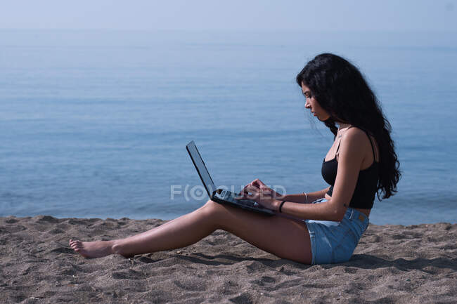 Приваблива жінка обробляє свій комп'ютер, сидячи на піску в — стокове фото