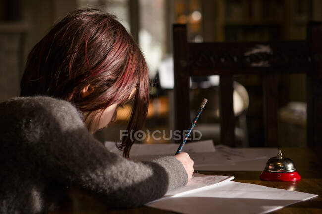 Little girl with bright red hair sits in patch of light writing story — Stock Photo
