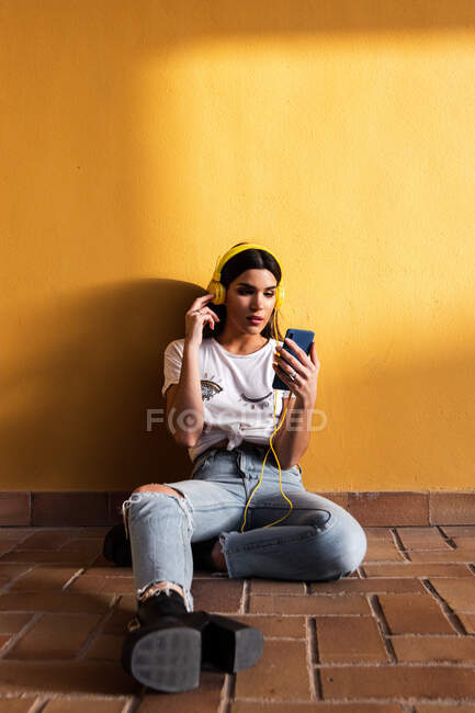 Portrait de fille brune espagnole assise sur le sol et écoutant de la musique avec écouteurs sur fond jaune mur. — Photo de stock