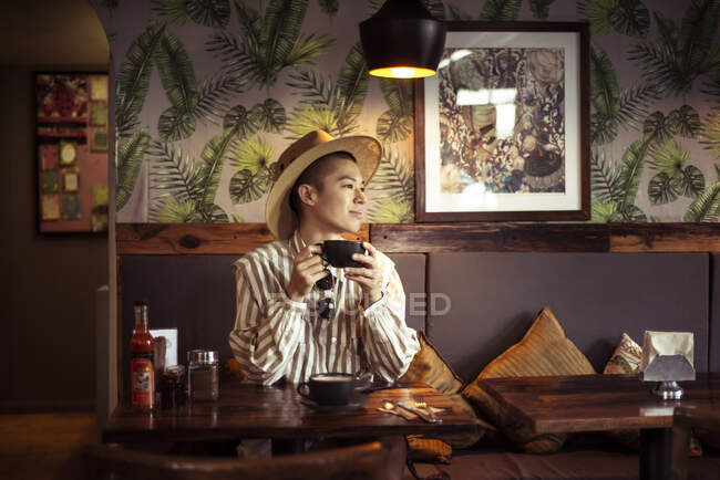 Alternative androgynous young traveller in hat drinks coffee at cafe — Stock Photo