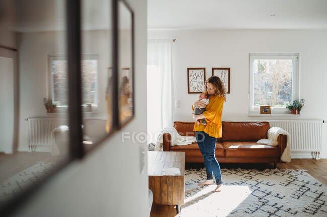 Mãe segurando bebê de pé na sala de estar com reflexão sobre fotos — Fotografia de Stock