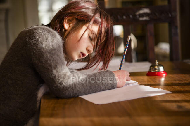 Focused little girl leans over paper writing with pencil at table — Stock Photo