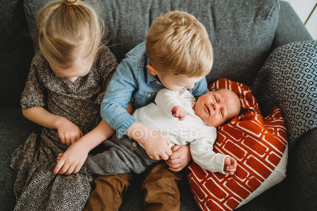 Bébé sur le canapé à la maison avec frère et sœur le tenant — Photo de stock