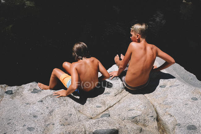 Dos chicos bronceados se sientan en un borde de roca sobre una oscura piscina de agua - foto de stock