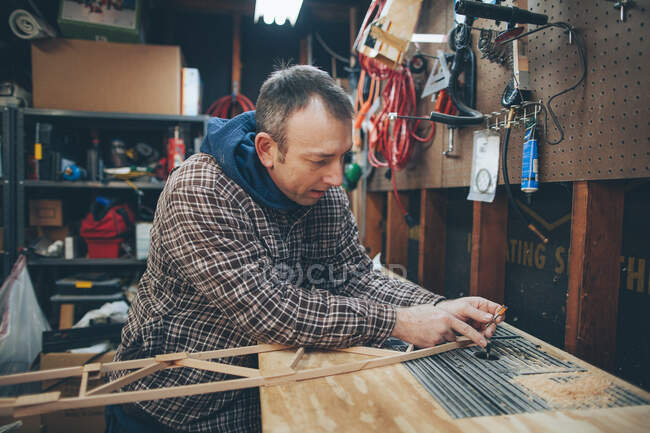Un hombre caucásico de mediana edad trabaja en una pequeña pieza de un avión de madera en su garaje. - foto de stock