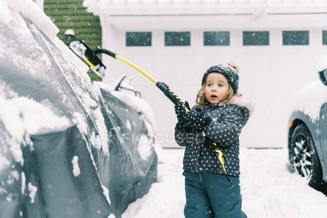 Маленькая милая малышка в снежном костюме очищает машины от снега кисточкой — стоковое фото