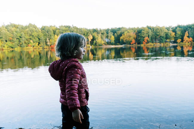 A little girl standing by a lake with colorful trees during autumn — Stock Photo