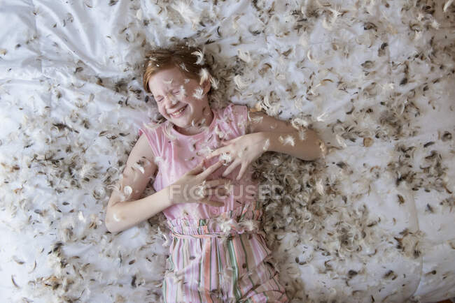 Chica pelirroja joven acostada en la cama durante una pelea de almohadas de plumas. - foto de stock