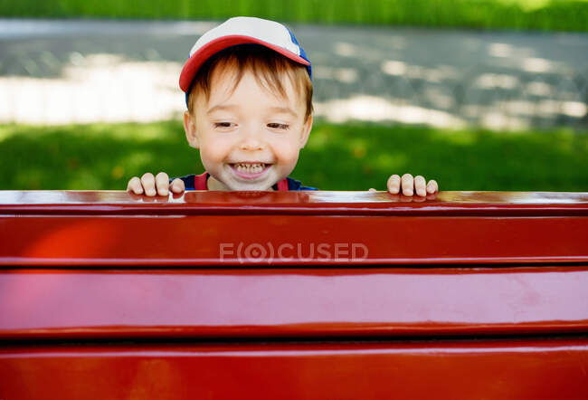 Lindo niño sonriendo felizmente asomándose desde detrás del banco del parque rojo - foto de stock