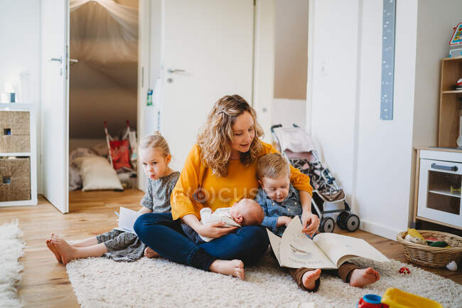 Mãe branca abraçando crianças lendo livros sentados no tapete em casa — Fotografia de Stock
