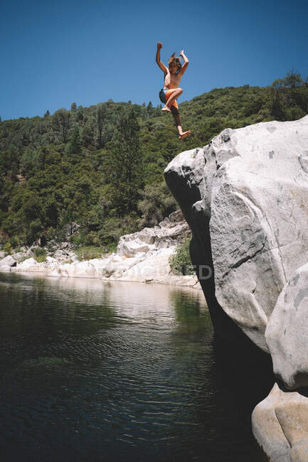 Menino salta com braços selvagens fora da rocha para o rio abaixo — Fotografia de Stock