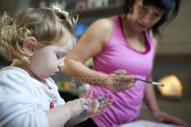 Petite fille mignonne regardant ses mains peintes avec sa mère en arrière-plan — Photo de stock