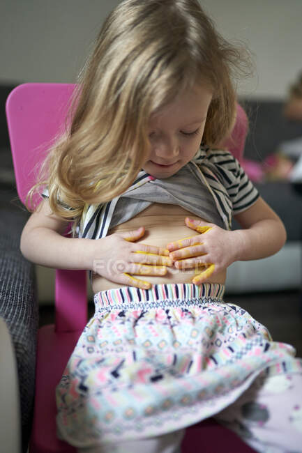 Cute happy little girl artist printing yellow painted palms on her belly. — Stock Photo