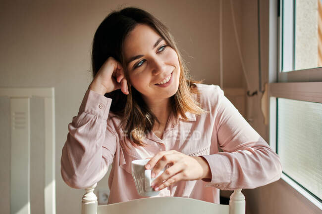 Femme sourit pendant qu'elle boit du café et regarde par la fenêtre — Photo de stock