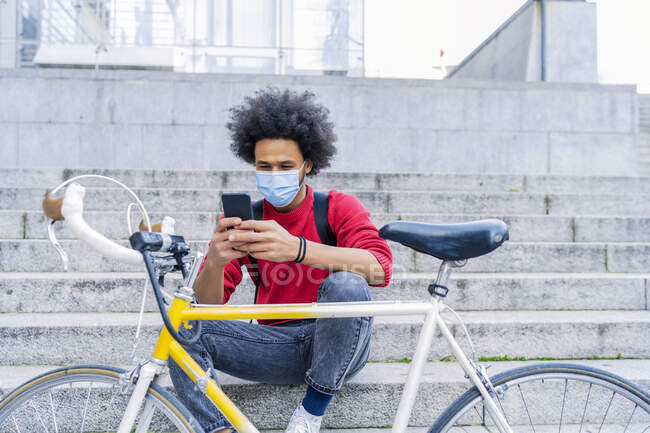 Young man with afro hair sitting sending a message next to his old bicycle — Stock Photo