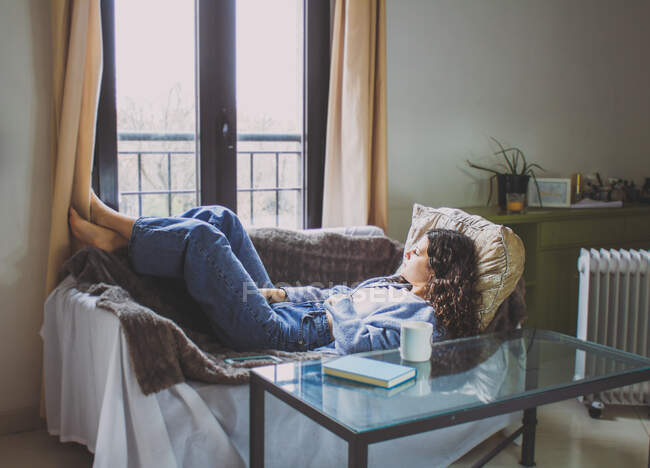 Молодая женщина лежит на диване и смотрит в окно с вьющимися волосами. — стоковое фото