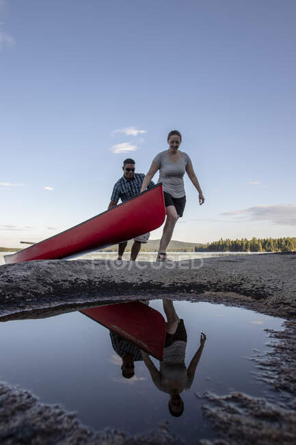 Hombre y mujer reflejados en charco acarreo canoa roja fuera del lago en Maine - foto de stock