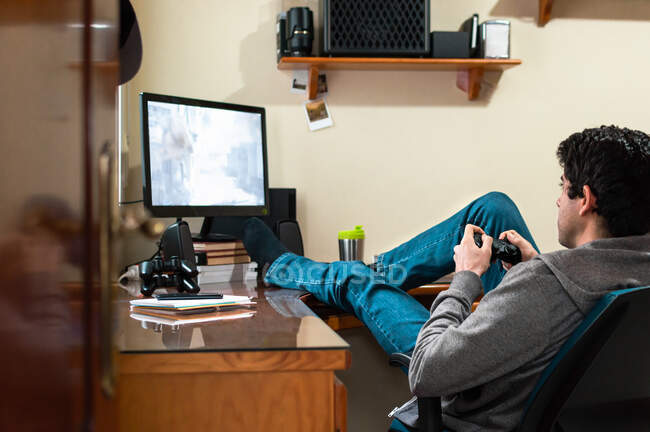 Молодой человек в толстовке и джинсах расслабляется, играя в видеоигры в своей спальне. — стоковое фото